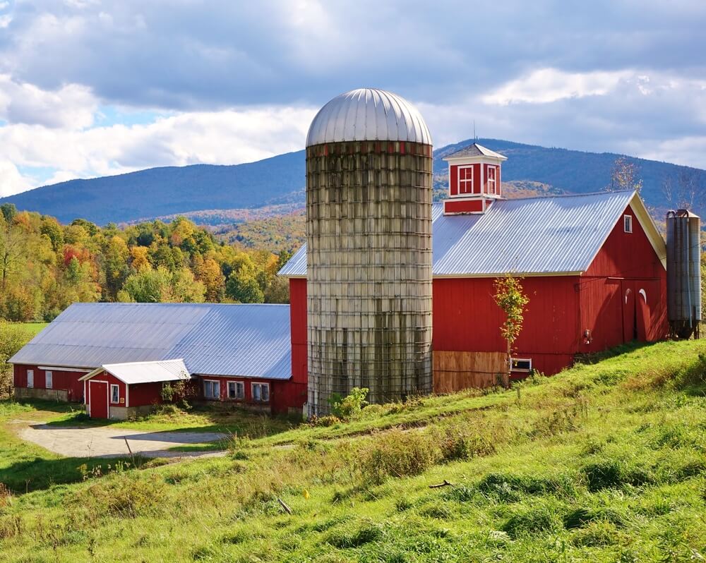 Red barn and silo with a sloping hill and green grass in foreground, trees with fall colors and Vermont mountains in the background under a blue and cloudy sky on a nice autumn day.